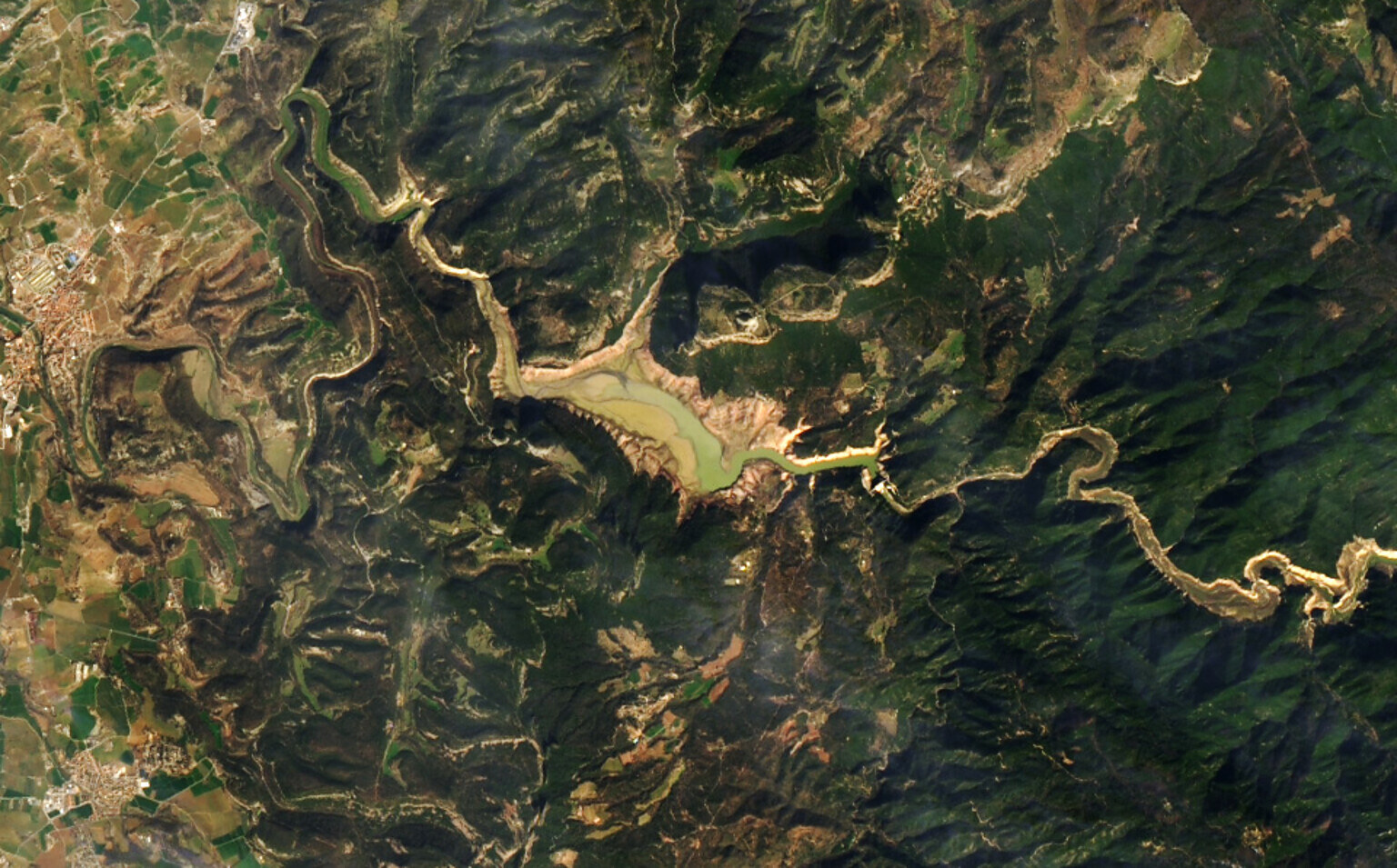 Satellite view of a European reservoir drained by drought — the shrunken water body sits as an exposed tan and olive basin where deep blue water would normally stand, with the river reduced to a thin thread through the surrounding hills